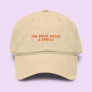 May include: A beige baseball cap with the text "THE BRIDE NEEDS A SPRITZ" in orange letters. The cap has a curved brim and a button on top. The background is a light purple color.