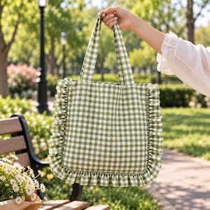 May include: A tote bag featuring a green and white gingham check pattern. The bag has two matching fabric handles and is decorated with a ruffled trim along the two side edges and the bottom edge, creating a gathered texture around the perimeter.