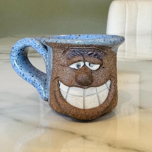 May include: A handmade ceramic mug with a smiling face design. The mug features a brown face with a wide, white-toothed grin, blue eyes, and a blue speckled handle. The rim and handle are a speckled blue.