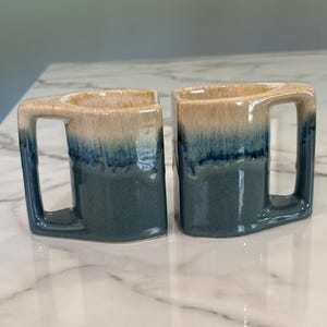 May include: Two ceramic mugs with a square-shaped handle design. Each mug features a two-tone glaze with a creamy beige top that transitions into a deep, dark blue base. The mugs have a smooth, glossy finish and a distinct, angular silhouette.