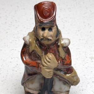 May include: A ceramic figurine of a soldier in a red coat, black boots, and a tall hat. The figurine has a mustache and is holding a rifle. The figure is painted with brown, tan, and white accents.