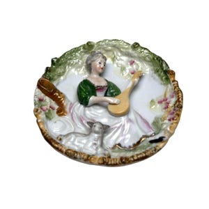 May include: A decorative porcelain plate featuring a woman in a green and white dress playing a lute. A lamb rests nearby. The plate has a scalloped edge with gold trim and floral accents. The background is white with green and pink details.
