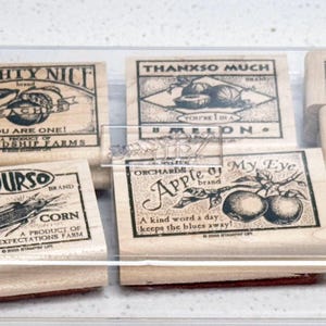 May include: A set of wooden stamps featuring fruit and vegetable designs and text. Stamps include "Mighty Nice Peaches", "Thankso Much Melon", "Yours Sweet Corn", and "Apple of My Eye". The stamps are in a clear plastic container.