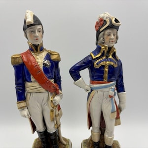 Op de afbeelding: Twee porseleinen beeldjes van militaire officieren in 18e-eeuwse uniformen. De figuren dragen blauwe jasjes met gouden epauletten, witte broeken en zwarte laarzen. Eén figuur draagt een rode sjerp, de andere een sjerp in de kleuren van de driekleur.