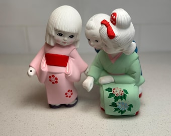 May include: Three ceramic Japanese dolls in traditional attire. The doll on the left wears a pink kimono with red floral accents and a red obi. The doll on the right wears a green kimono with floral designs and a red bow. The middle doll is wearing a blue kimono.