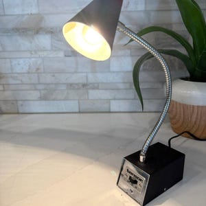 May include: A vintage Tensor desk lamp with a black base and a flexible, silver-toned neck. The lamp has a conical, gray lampshade and a switch labeled "OFF LO HI". The lamp is turned on, emitting a warm, yellow light.