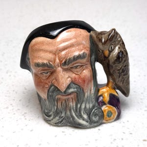 Könnte beinhalten: Eine Keramiktasse in Form eines bärtigen Männerkopfes mit schwarzem Hut und einer Eule an der Seite. Die Tasse weist detaillierte Gesichtszüge auf und ist in Braun-, Grau- und Schwarztönen bemalt.