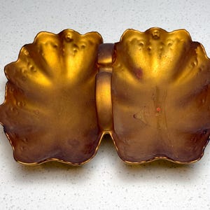 May include: A decorative, two-sectioned dish with a handle, featuring a golden-bronze finish. The dish has a scalloped edge and a textured surface, adding visual interest. The handle connects the two sections.