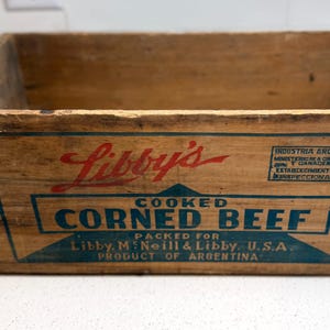Könnte beinhalten: Eine Vintage-Holzkiste mit dem Libby's-Logo in Rot und den Worten "Cooked Corned Beef" in Blau. Die Kiste ist aus hellem Holz gefertigt und trägt den blauen Text "Packed for Libby, M'Neill & Libby, U.S.A. Product of Argentina."