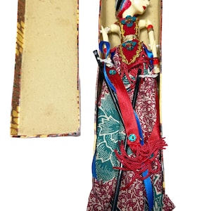 May include: A Wayang Golek puppet with a pale face, dark hair, and a red bodice featuring gold trim. The puppet wears a long skirt with a green and maroon floral batik pattern, accented by blue ribbons, red tassels, and two black control rods.