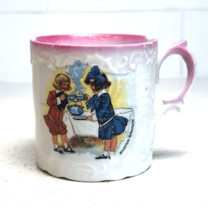 May include: White ceramic mug with a pink rim and handle, featuring a Buster Brown illustration. The artwork depicts two children serving tea. The mug has a decorative, scalloped edge.