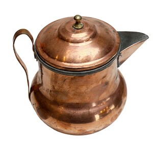 May include: A copper pitcher with a rounded body, a curved handle, and a fitted lid featuring a small brass-colored knob. The pitcher has a wide spout and a dark metallic rim around the top edge, showing a polished yet weathered surface.