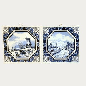 Op de afbeelding: Twee blauw-witte keramische tegels met Hollandse landschappen. Eén tegel toont een windmolen, de andere een kanaalscène. Elke tegel heeft een decoratieve blauwe rand met bloemen- en geometrische patronen.
