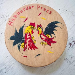 Puede incluir: Prensa de hamburguesas redonda de madera con un diseño de gallo pintado. Los gallos son amarillos y rojos con alas negras. Las palabras "Hamburger Press" están en rojo, arqueadas en la parte superior.