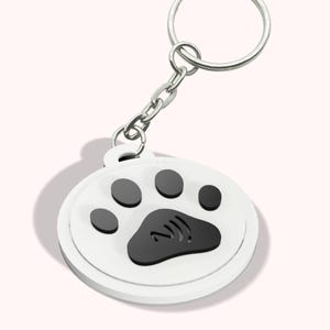 Puede incluir: Llavero blanco y negro con huella de pata, con cadena y anilla plateadas. El llavero tiene el texto "NFC KEYCHAIN FILE" en la parte inferior. El diseño de la huella de pata es un accesorio bonito y sencillo.