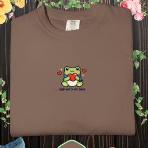 Peut inclure: Sweat-shirt couleur expresso à col rond. Le t-shirt présente une grenouille de dessin animé brodée tenant un cœur, entourée de cœurs, avec le texte "HOP INTO MY DMS" en dessous. Le t-shirt est fait d'une matière douce.
