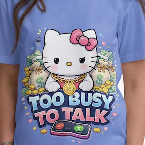 May include: A light blue t-shirt featuring a cartoon cat with a pink bow, gold chain, and the text "TOO BUSY TO TALK". The cat is surrounded by money bags and coins, with a smartphone graphic below.