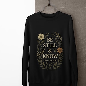 May include: Black sweatshirt with the text "BE STILL & KNOW THAT I AM GOD" in a floral wreath design. The design includes beige and brown flowers and green leaves. The sweatshirt is hanging on a metal hanger.