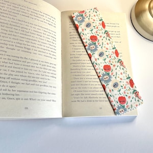 Op de afbeelding: Een stoffen bladwijzer met blauwe en rode bloemmotieven, geplaatst in een open boek. De bladwijzer is rechthoekig en heeft een herhalend ontwerp. Een lamp is zichtbaar op de achtergrond.