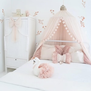 May include: A white bed with a pink canopy, pillows, and a swan-shaped plush toy. The canopy is draped over the bed, and the pillows are arranged against the headboard. A white wardrobe with a large pink bow is in the background.