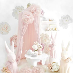 May include: A whimsical nursery scene with a pink canopy, white table, and decorative bunny statues. The room is adorned with large paper flowers and a tea set, creating a soft, inviting atmosphere. The color palette is primarily soft pinks and whites.