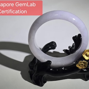 Könnte beinhalten: Ein helllavendelfarbenes Jade-Armreif-Armband, das auf einem schwarzen Holzständer mit goldenen Akzenten ausgestellt ist. Der Text "Singapore GemLab Certification" befindet sich in einem roten Feld oben links. Das Armband ist rund und glatt.