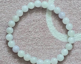 May include: A bracelet made of round, light-colored beads. The beads are primarily white with a few pale lavender beads interspersed. The bracelet is resting on a pink, textured surface.