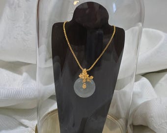 May include: A gold necklace with a circular, pale green pendant, displayed on a black stand inside a glass cloche. The pendant is adorned with gold filigree. The necklace is presented on a white, patterned fabric.