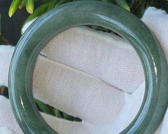 May include: A smooth, jade green bangle bracelet. The circular bracelet is held in a gloved hand, with green foliage in the background. The jade is a solid color with a glossy finish. A classic jewelry piece.