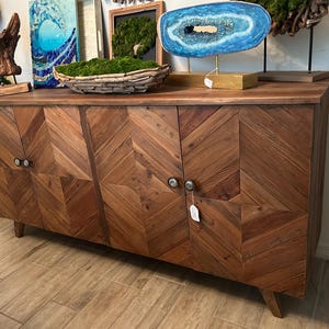 Op de afbeelding: Een houten kast met vier deuren, elk met een chevron-patroon. De kast heeft een donkerbruine afwerking en zilveren deurknoppen. De kast staat op een houten vloer.