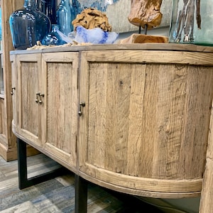 May include: A curved wooden cabinet with three doors and black metal legs. The cabinet has a rustic, weathered look and is made of reclaimed wood.