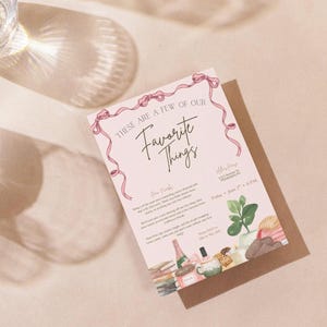 May include: A light pink invitation card with the text "These Are A Few of Our Favorite Things" and pink ribbon decorations. The card features illustrations of a plant, books, a bottle of champagne, and a teacup. The card is on a brown envelope.