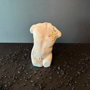 Puede incluir: Una estatua de torso esculpido de color crema con detalles dorados. La estatua es una representación estilizada de la forma humana, con detalles visibles de la espalda y los hombros. La escultura está colocada sobre una superficie oscura salpicada de pequeñas piedras oscuras.