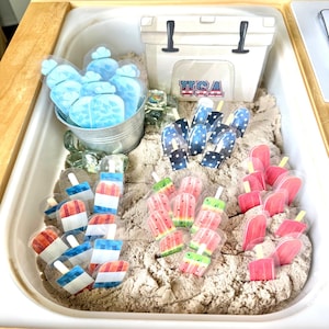 May include: A sensory bin filled with sand, featuring various popsicle-shaped cutouts in red, white, and blue, along with watermelon and pink popsicles. A white cooler with "USA" text and a metal bucket with blue ice shapes are also present.