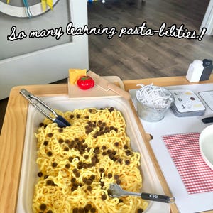Puede incluir: Una mesa de juego de madera con una bandeja blanca llena de espaguetis amarillos y bolas marrones. Un tomate de juguete, queso y un cuchillo de madera están sobre una tabla de cortar. El texto "So many learning pasta-bilities!" está en la parte superior.