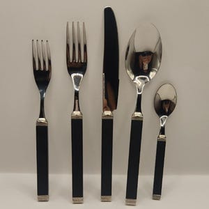May include: A set of five pieces of silverware, including a fork, knife, spoon, and two smaller spoons. The silverware has black handles with silver accents and polished metal heads.