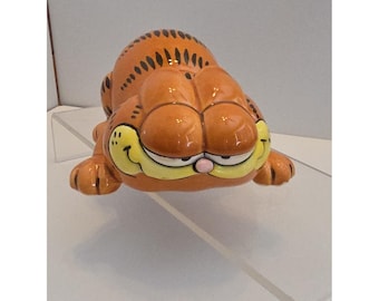 Puede incluir: Una figura de cerámica del gato atigrado naranja Garfield, representado en una posición tumbada y de gateo. La figura presenta rayas negras, un hocico amarillo, una pequeña nariz rosa y una amplia sonrisa con grandes ojos entrecerrados.