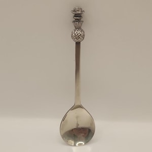 May include: A silver-colored spoon with a pineapple and hat design on the handle. The spoon has a rounded bowl and a long, slender handle. The pineapple is textured, and the hat is detailed.