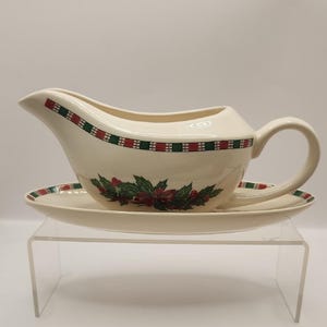 May include: Cream-colored gravy boat and saucer set with a festive Christmas design. The boat features a handle and spout, with a red and green plaid border and holly and berry accents. The saucer mirrors the design.
