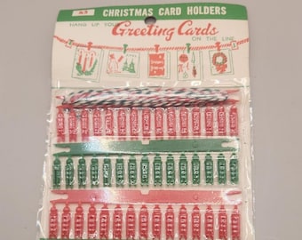 Op de afbeelding: Kerstkaarthouders in een doorzichtige plastic verpakking. De verpakking heeft een groen-witte koptekst met de tekst "CHRISTMAS CARD HOLDERS" en "Greeting Cards". De houders zijn rood en groen, met een koord om op te hangen.