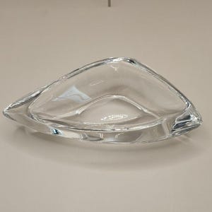 May include: A clear glass decorative dish with a unique, asymmetrical shape. The dish has a smooth, curved design and is made of transparent glass, allowing light to pass through. The dish is approximately 10 inches long.