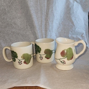 May include: Three ceramic pieces: two mugs and a creamer. Each piece is off-white with a green and red leaf and berry design. The mugs have handles, and the creamer has a spout and pedestal base. The design is repeated on each piece.
