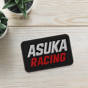 May include: Black rectangular patch with the text "ASUKA RACING" in white and red. The patch is on a light wood surface, next to two small potted succulents.