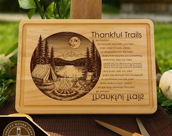 Op de afbeelding: Rechthoekige houten snijplank met een gegraveerd ontwerp. Het ontwerp toont een kampeerscène met een tent, kampvuur en meer, samen met de tekst "Thankful Trails". Gemaakt van lichtgekleurd hout.
