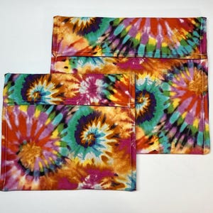 Può includere: Due pochette in tessuto tie-dye nei toni dell'arancione, giallo, rosa, viola e verde acqua. Le pochette hanno una forma rettangolare con chiusura a patta. Il tessuto presenta un motivo a spirale.