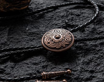 May include: A copper-colored bolo tie with a detailed floral design on the circular pendant. The tie features a braided black leather cord and decorative metal tips. The bolo tie rests on a dark, textured surface.