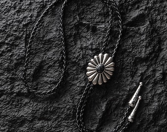 May include: A black braided leather bolo tie with a silver-toned daisy-shaped pendant featuring a black stone center. The tie has decorative silver tips. The bolo tie is displayed on a textured black surface.