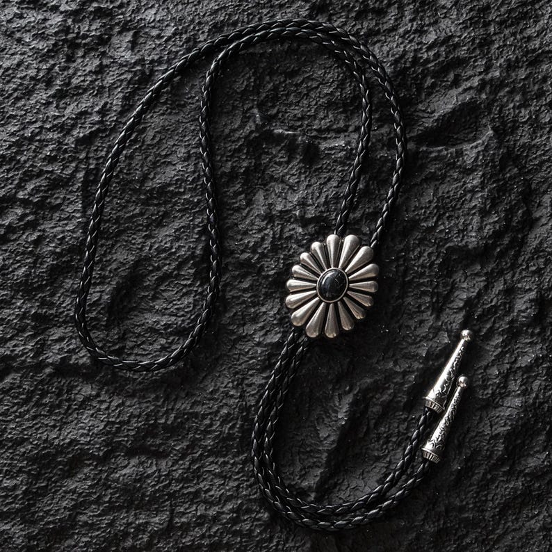 May include: A black braided leather bolo tie with a silver-toned daisy-shaped pendant featuring a black stone center. The tie has decorative silver tips. The bolo tie is displayed on a textured black surface.