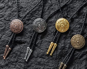 May include: Close-up of four bolo ties with decorative metal medallions. The medallions are in copper, silver, and gold tones, each with an embossed floral design. The ties have black braided cords and matching metal tips.