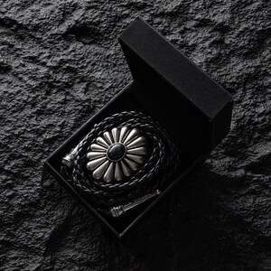 May include: A black bolo tie with a silver-colored floral design and a black stone center, presented in a black box. The braided leather cord is coiled within the box, with the silver-toned tips visible.
